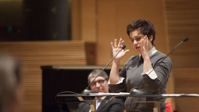 Silvia Modig står vid ett podium och gestikulerar i riksdagen.