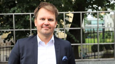En man med ljusbrunt hår iklädd en svart kostym står utomhus med staket och träd i bakgrunden.