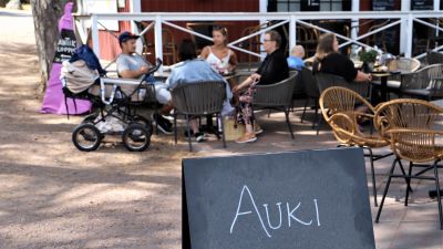 En familj med en barnvagn sitter på en uteservering i Nagu gästhamn. I förgrunden en skylt med texten "Auki".