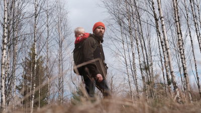 Ulkoiluvaatteisiin pukeutunut mies pieni lapsi selässään katsoo miettiväisenä kaukaisuuteen syksyisessä koivikossa