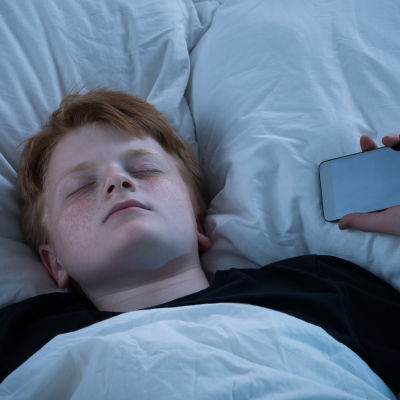 Pojke som somnat i sängen med mobiltelefon i ena handen.