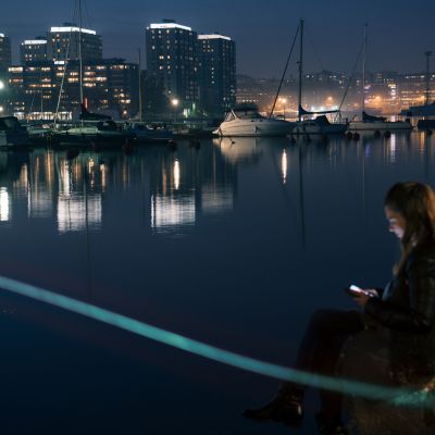 Tyttö istuu illalla satamassa veden äärellä, käyttää puhelintaan