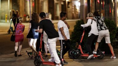 En person åker elsparkcykel på trottoar på natten. Två personer bakom honom åker på samma elsparkcykel.