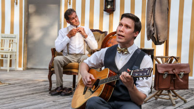 Spelamannen spelar gitarr i förgunden. Bakom honom sitter Agaton och knäpper översta knappen på sin skjorta. De är klädda i gråvita kostymkläder.