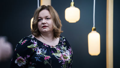 Krista Kiuru i blommig skjorta står framför en mörk vägg. I bakgrunden syns också lampor. Kiuru tittar åt sidan.