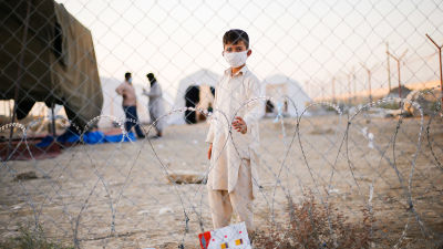 En liten pojke i munskydd tittar in i kameran genom stängsel på gränsen mellan Afghanistan och Iran. I bakgrunden syns tält och två män som pratar med varandra.