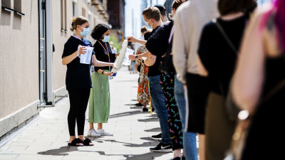 Emmi Sundén och Lura Aunimo delade ut vatten till personer som väntar i vaccinationskön. På bilden tar en ung man en pappmugg från en stor mugghögg som en kvinna håller i. En kvinna står bredvid med en vattenkanna i handen redo att fylla koppen.