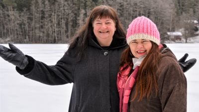 Två kvinnor står i ett snöigt landskap och tittar leende in i kameran. Den ena kvinnan har händerna utbredda i en posering.