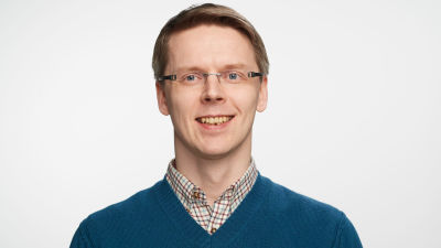 Lyhyttukkainen, silmälasipäinen mies katsoo kohti ja hymyilee, lähikuva.