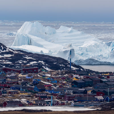 Ilmakuvassa näkyy grönlantilainen rannikkokylä sekä sen takana jäätikköä.