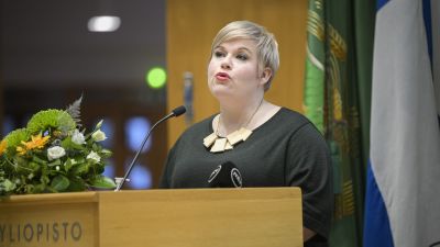 En kvinna i grön tröja håller tal vid ett podium