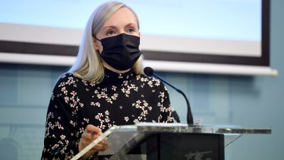 Maria Ohisalo bär en blommig, svart klänning och ett svart munskydd och står vid ett talarpodium.