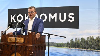 Petteri Orpo står och håller tal vid partimötet i Seinäjoki, bakom sig har han ett sjölandskap.