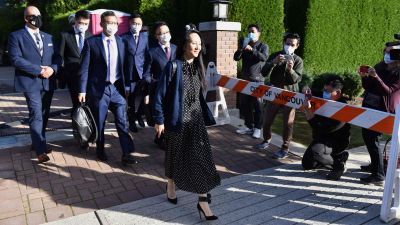 Kvinna i klänning går utomhus i soligt väder med en grupp kostymklädda män i munskydd. Huaweis finanschef Meng Wanzhou lämnar sitt hem i Vancouver den 24 september 2021.