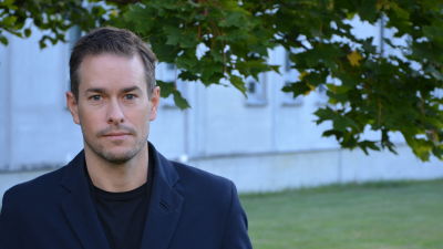 Kim Strandberg, en vit man, står framför ett träd och ett betonghus.