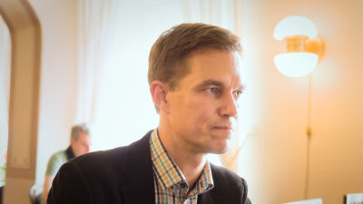en man i kort ljusbrunt hår och svart kavaj står inomhus i ett ljust rum