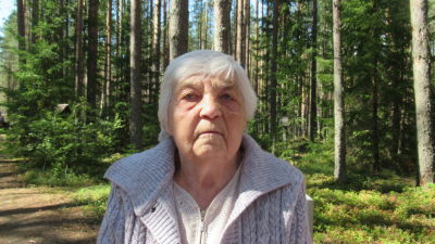 En äldre dam tittar rakt in i kameran. Bakom henne tät skog.