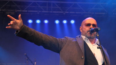 Isländska artisten Bubbi Morthens sjunger i mikrofon, med handen utsträckt, mot blå bakgrund