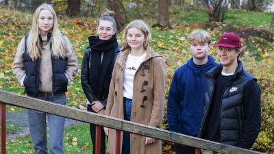 Fem ungdomar står på rad utomhus, de tittar in i kameran.