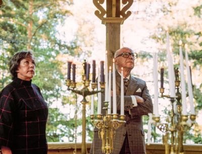 Tekstiilitaiteilija Armi Ratia ja presidentti Urho Kekkonen ihastelevat kynttiläkruunuja verannalla kesäisessä maisemassa. Kuva Bökarsin kartanosta Porvoosta.