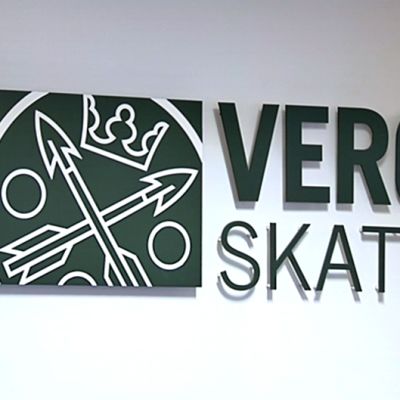 Vero- ja skatt-tekstit logosymbolin kanssa valkoisella seinällä. Tekstit ovat hieman irti seinästä, jolloin niiden alle muodostuu pienoinen varjo.