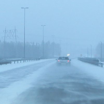 Auto ajaa lumisateessa ja tuulessa.