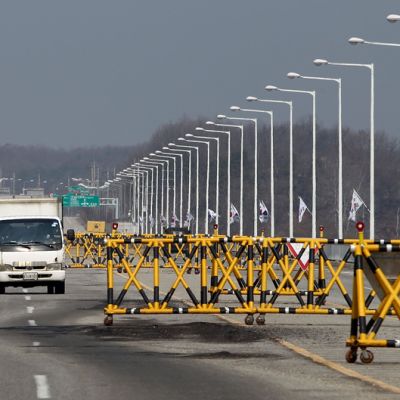 Kuvassa kaksi kulkuneuvoa ajaa keltamustien porttien välistä, ajotien vierellä liehuu Etelä-Korean lippuja.