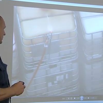 Pelastustoimen päivystävä päällikkö Pauli Nurminen näyttää tiedotustilaisuudessa pienoiskopterista kuvattua videota räjähdekonttien rivistöstä. Kuvassa näkyy konttiin asennettu vesiletku, jonka kautta konttiin pumpattiin vettä sen lämpötilan alentamiseksi.