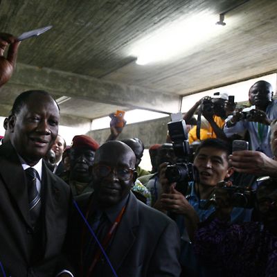 Alassane Ouattara pitää äänestyslippua koholla edessään olevan vaaliuurnan (ei näy kuvassa) yläpuolella. Tiedotusvälineiden edustajat kuvaavat tapahtumaa.