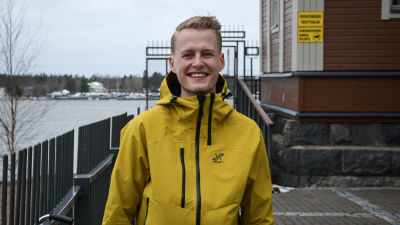 Ung man i gul jacka står utomhus, tittar leendes in i kameran. I bakgrunden syns vatten.