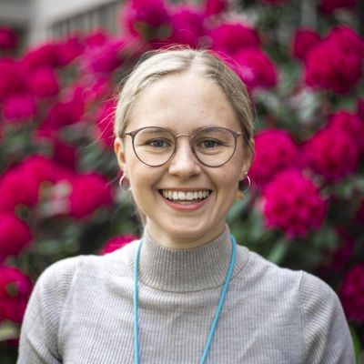 En unga kvinna ler och ser in i kameran. Hon har på sig glasögon. I bakgrunden syns röda blommor.