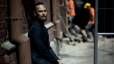 Ilmari Käihkö. En svartklädd man lutar mot en tegelvägg. Bilden är mörk och han har en allvarlig min. Mannen har kort ljusbrunt hår och skägg.
