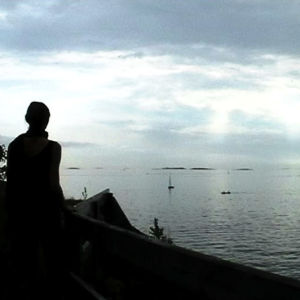 Nainen meren rannalla katsoo poispäin kamerasta, kohti horisonttia.