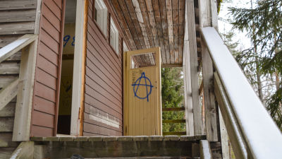 En rödbrun träbyggnad i dåligt skick. En dörröppning saknar dörr, den andra har en dörr som står öppen. Någon har ritat en anarkistsymbol på dörren.