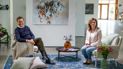 Mika Ihamuotila sitter i en fåtölj till vänster i ett vardagsrum. I en fåtölj till höger sitter Ira Hammermann. Båda ser leende in i kameran.