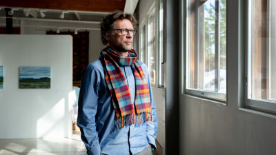 Antti står i ett soligt konstgalleri och ser ut genom fönstret. Han har en blå skjorta och röd-blå-rutig halsduk på sig.