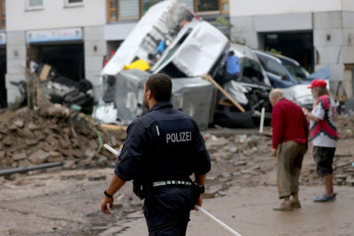 En polis går på en lerig väg i en stadsskärna. Framför honom finns en hög med flera bilar. I bakgrunden syns ett äldre par. Mannen är framåtlutad och kvinnan håller sin hand på hans rygg.
