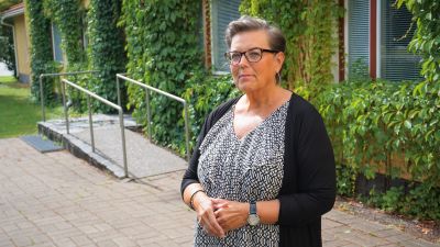 Kvinna med kort hår och glasögon står framför en gul byggnad.