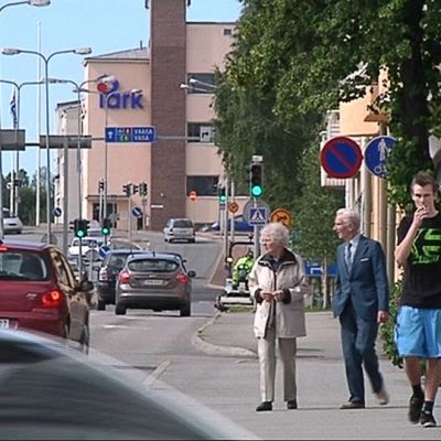 Kuvassa liikennettä Kokkolan keskustassa. Autoja tulee ja menee, liikennevalot ovat vihreällä. Jalkakäytävällä kävelee ihmisiä.