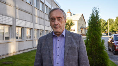Man i övre medelåldern iklädd skjorta och kavaj står utomhus och tittar in i kameran. I bakgrunden grön gräsmatta och fasaden av ett betonghus.