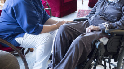 En skötare håller en äldre persons hand. Den äldre personen sitter i rullstol.