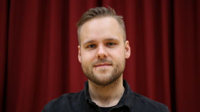 Mansperson står på teaterscen, i bakgrunden rött sammetsdraperi.