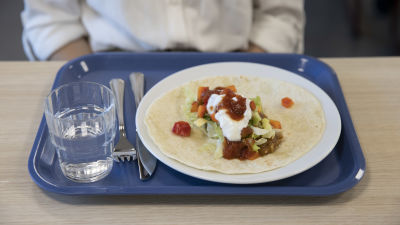 En tallrik med en tortilla med vegetarisk sås på. Under den en skolmatsalsbricka av blå plast.