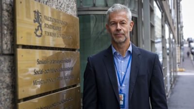 En gråhårig man står vid Social- och hälsovårdsministeriets byggnad. Han tittar in i kameran och ler något. Han är klädd i mörk kavaj och en blå skjorta.