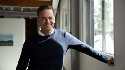 Mika Ihamuotila står vid ett fönster lutandes mot fönsterkarmen. Han ler rakt in i kameran.