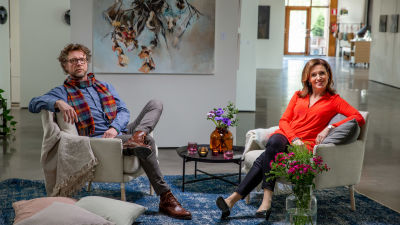 Antti Reini och programledaren Ira Hammermann sitter i varsin vit fåtölj i ett konstgalleri.