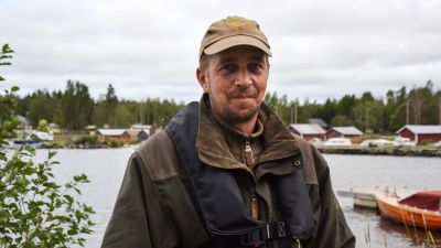 Man i jaktkläder och flytväst tittar in i kameran, i bakgrunden syns en småbåtshamn.