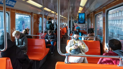 Passagerare sitter och står i en metrovagn med orangefärgade bänkar. Många eller alla ser ut att ha munskydd.