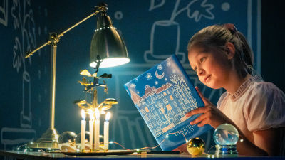 Jemina Haukka lukee Ihmeiden talo -kirjaa päydän äärellä. Pöydällä on lamppu, joulukoristeita sekä pieni lumisadepallo.
