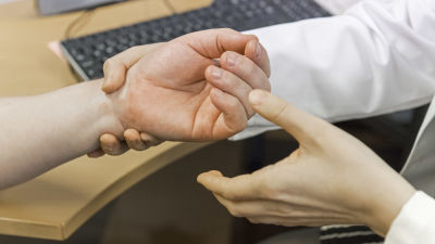 Ett par läkarhänder och en patients hand. I bakgrunden ett tangentbord.